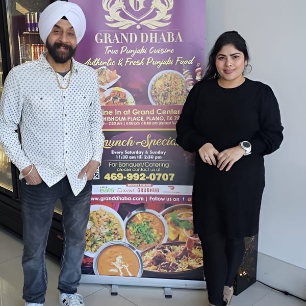 Two people stand beside a Grand Dhaba banner showcasing Indian dishes and restaurant information.