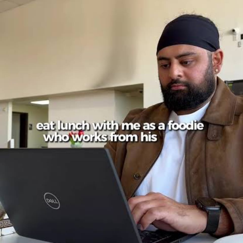 Bearded man wearing a head covering and brown jacket works on a Dell laptop indoors, with overlaid text about eating lunch.