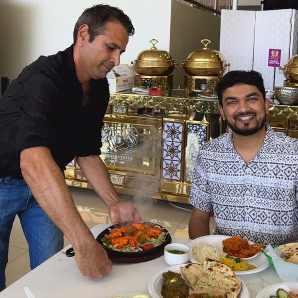 A Grand Dhaba server places a sizzling Indian chicken dish before a smiling customer.