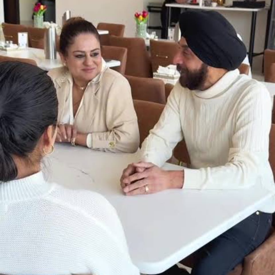 Three people converse around a white table in a warmly furnished restaurant dining room.